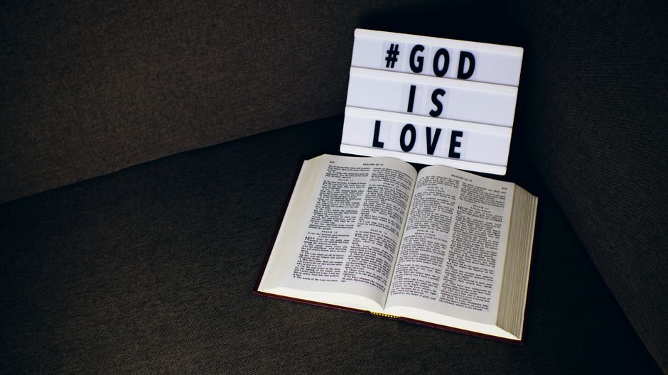 Open Bible placed beside a lightbox sign reading 'God is Love' on a dark fabric surface.